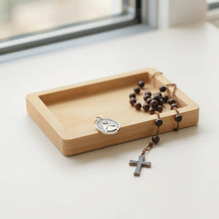 Metal saint charm pendant with a cross motif resting beside rosary beads on a light wooden tray